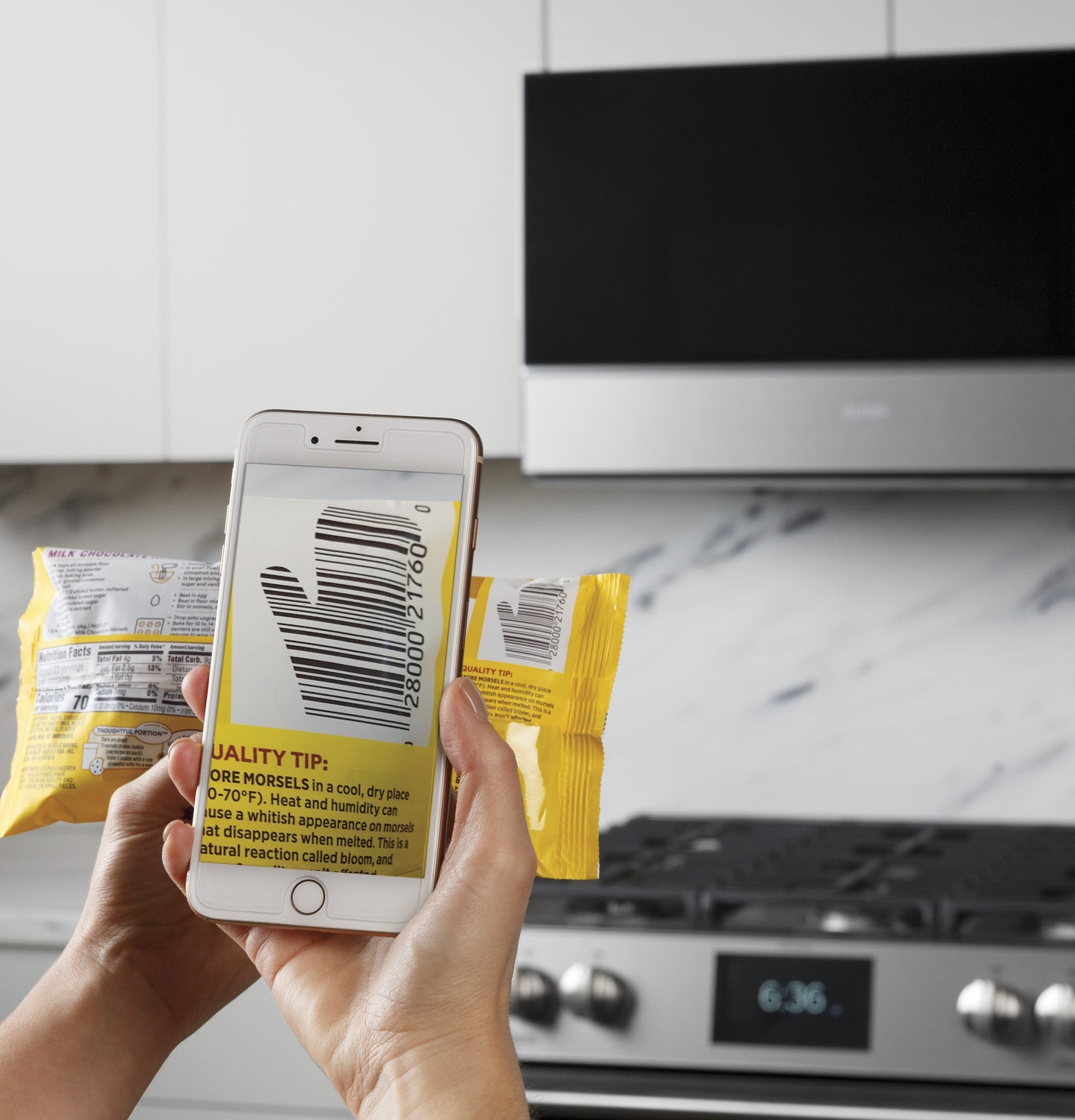 Person scanning a barcode on a yellow food package with a smartphone in front of a stainless steel stove and microwave in kitchen