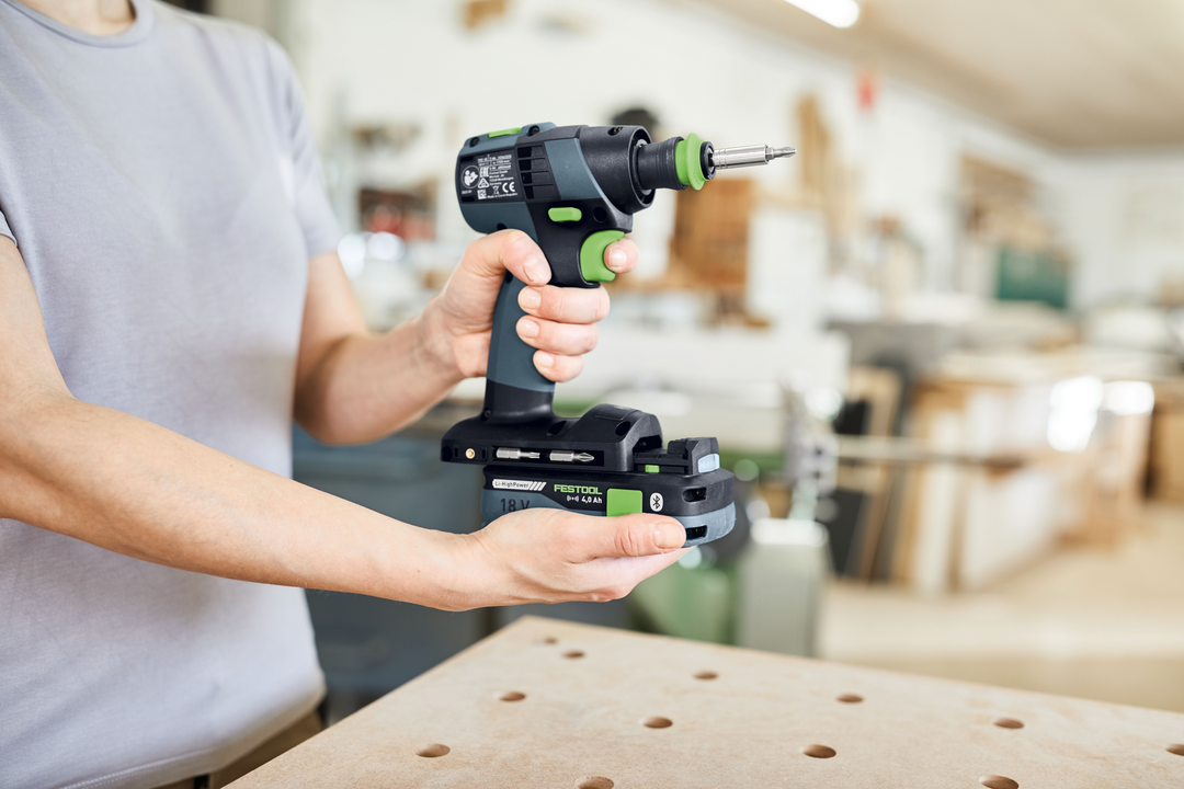 Person holding a compact Festool cordless drill in a woodworking workshop with tools and equipment in the background
