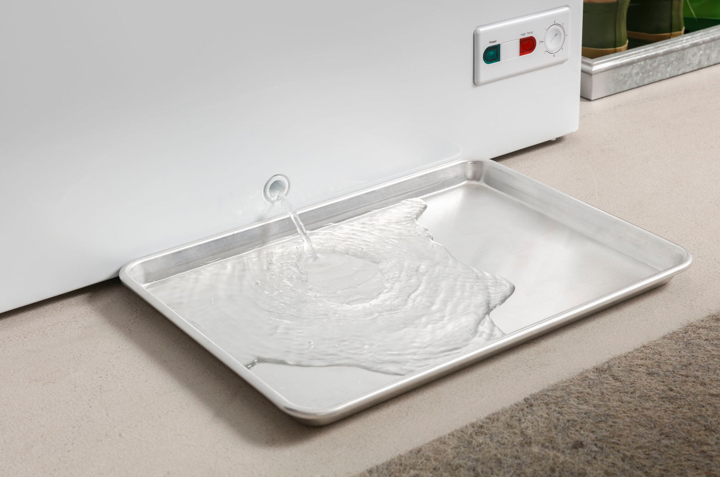Water draining from manual defrost chest freezer into removable metal drip tray on beige carpet flooring