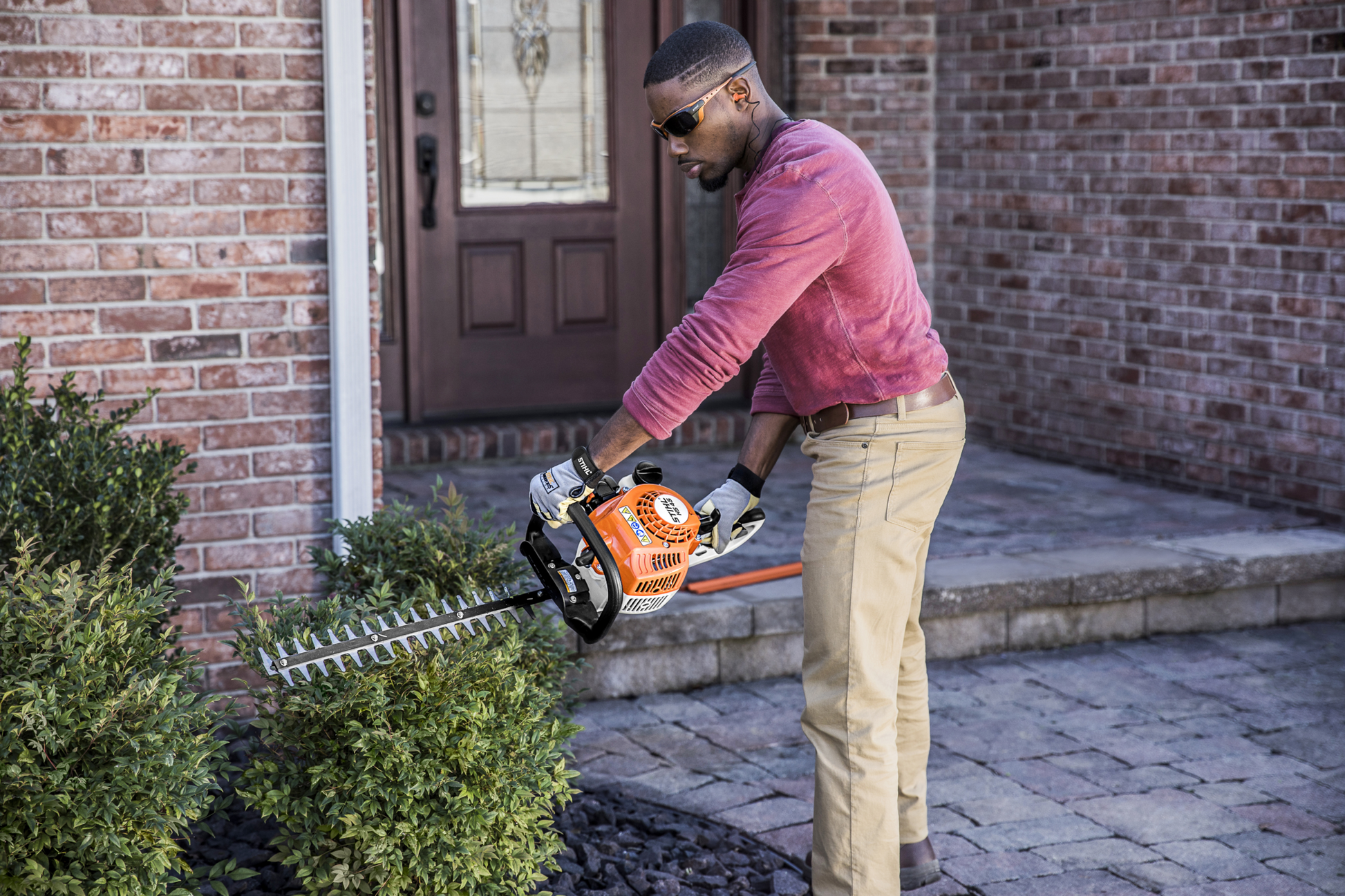 Person wearing gloves using lightweight orange and black hedge trimmer to shape outdoor shrubbery near brick house entryway