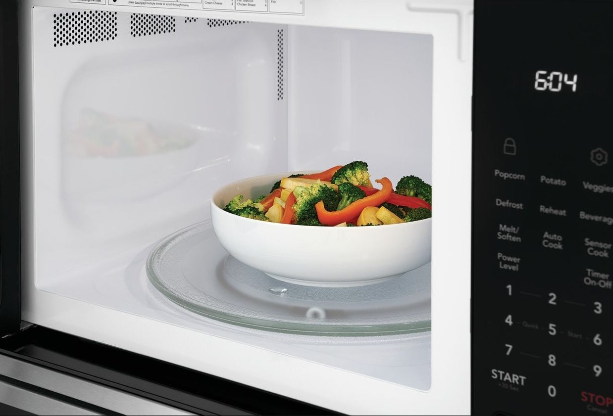 White microwave interior with a rotating glass turntable holding a bowl of mixed steamed vegetables and a digital control panel displaying time 6:04