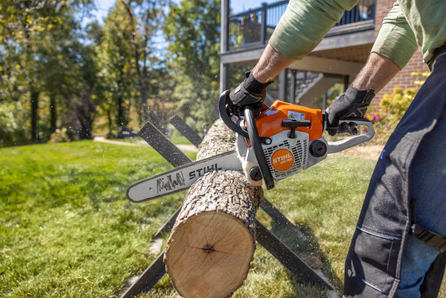 Person using an orange STIHL gas-powered chainsaw to cut a log outdoors on a sunny day in a backyard