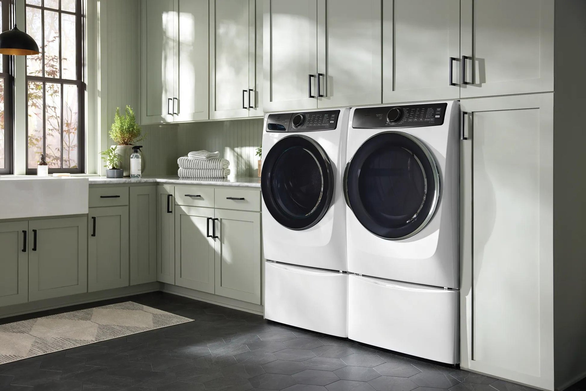 White front load washers with black doors and control panels installed in a light green cabinetry laundry room setting