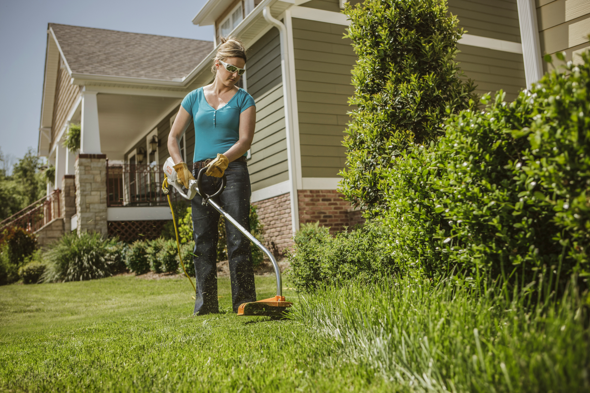 Woman using STIHL FSE 60 Electric Trimmer to cut tall grass along a landscaped yard near a suburban home