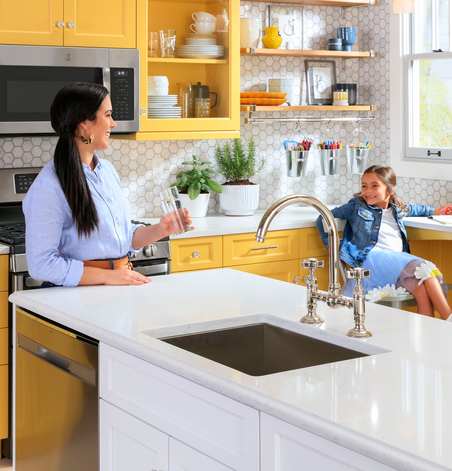 Bright kitchen with yellow cabinets, stainless steel appliances, white countertop, and woman talking to child by window seat