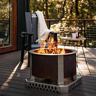 Smokeless corten steel fire pit with stainless steel legs burning on deck near chair and outdoor table setting