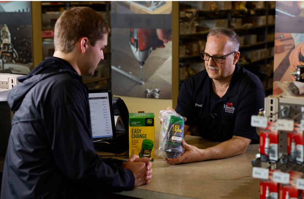 Store employee explaining a home improvement tool to a customer over a counter with product packaging and a computer screen visible.