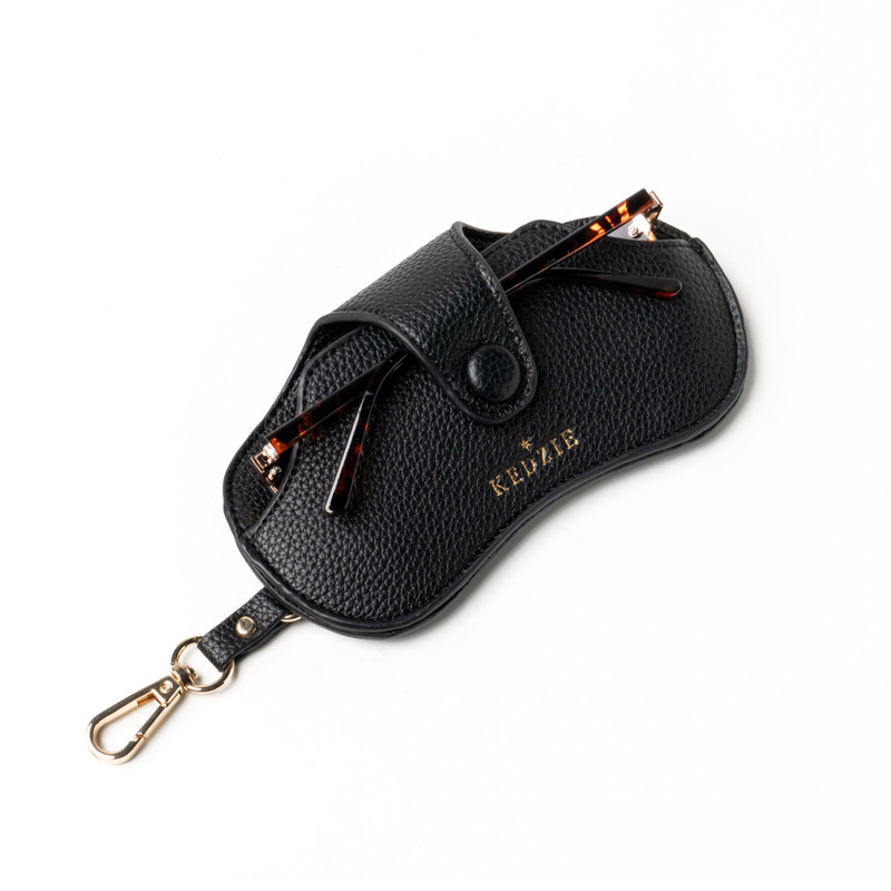 Black textured vegan leather eyewear case with gold embossed logo, snap closure, and gold clasp, shown with tortoiseshell glasses partly inserted, displayed on white background in Accessories and Bags & Wallets collections.