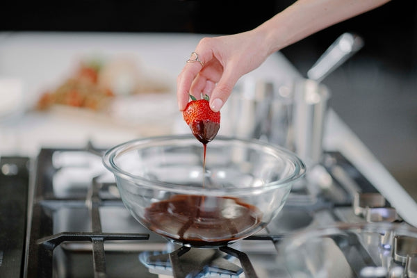 Close-up of a hand dipping a strawberry into a glass bowl with melted chocolate on a stainless steel gas cooktop