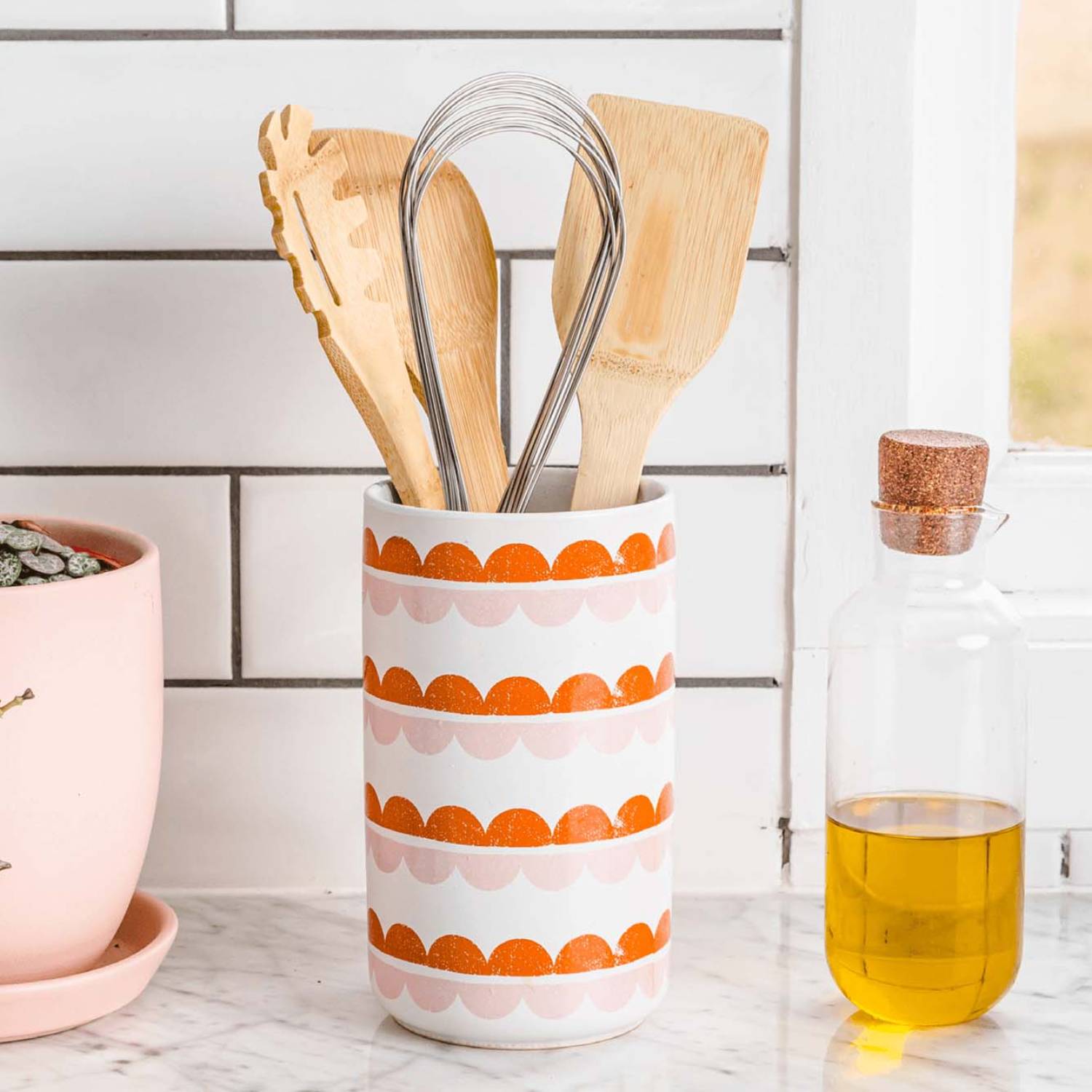 Red-handled stainless steel kitchen whisk alongside wooden utensils in decorative ceramic holder on countertop