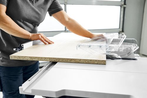 Person guiding a wood panel through a cordless Festool table saw with clear safety guard in a workshop setting