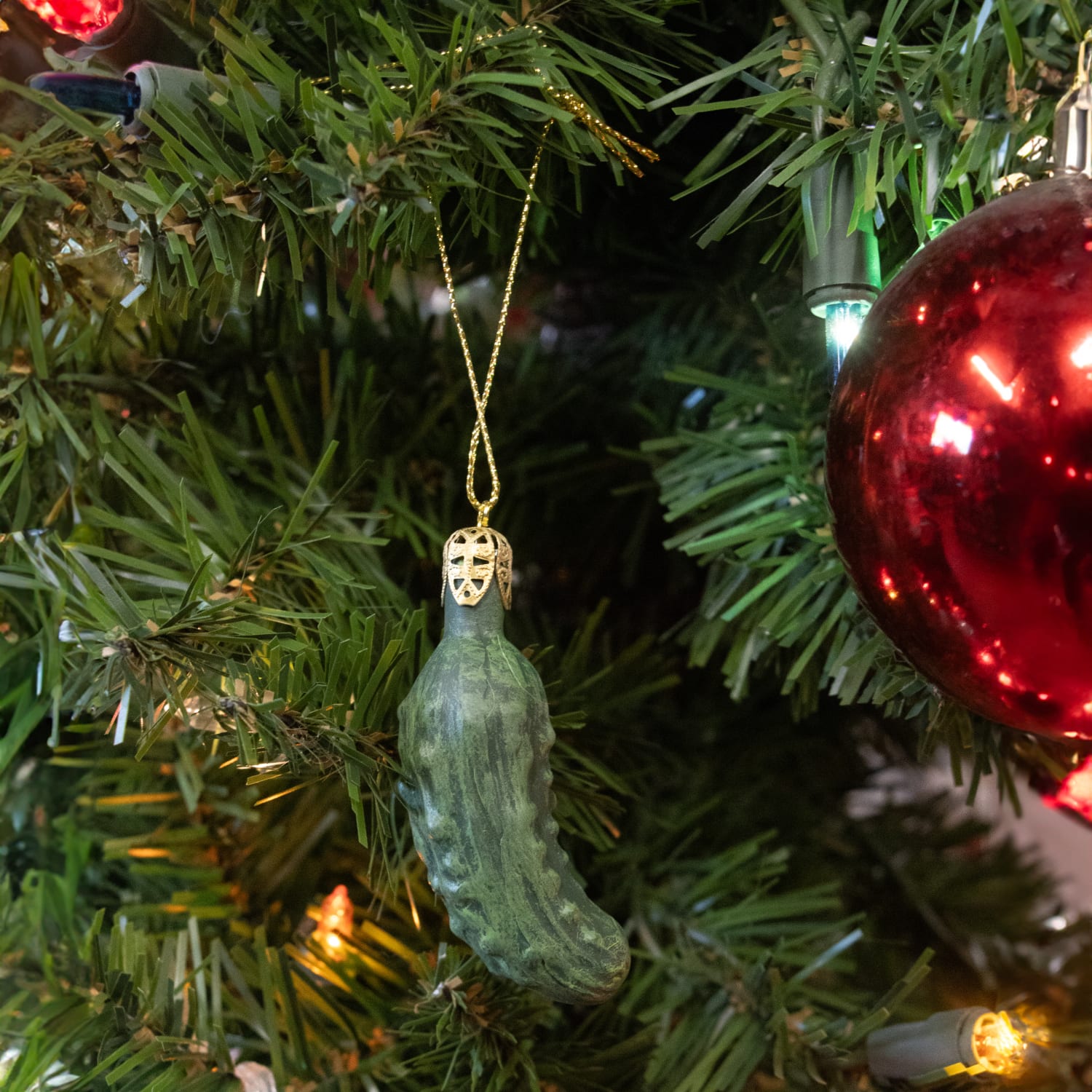 Green pickle-shaped ornament hanging from gold string on artificial tree with red bauble in Christmas collection