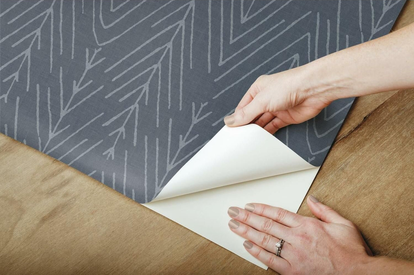Peel and stick wallpaper with navy background and subtle twig pattern being applied over wooden surface for home decor