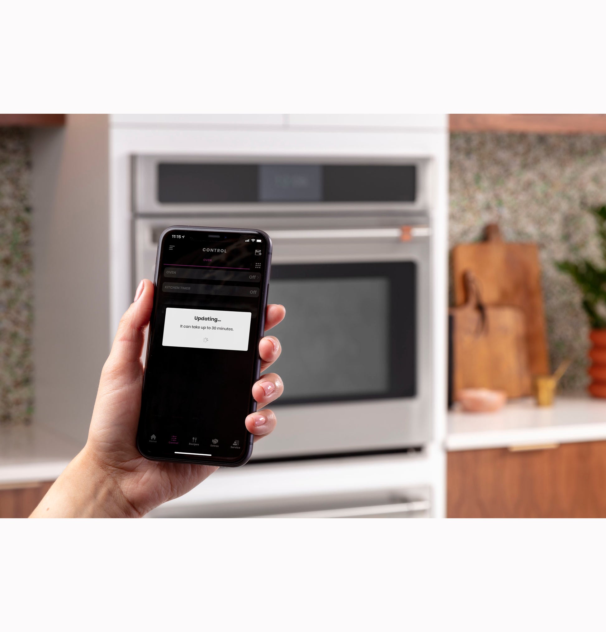 Hand holding smartphone showing updating screen near stainless steel wall oven in modern kitchen setting