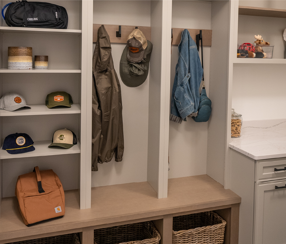Organized mudroom with built-in cubbies featuring hats, coats, a backpack, woven baskets below, and a white countertop with cabinetry on the side
