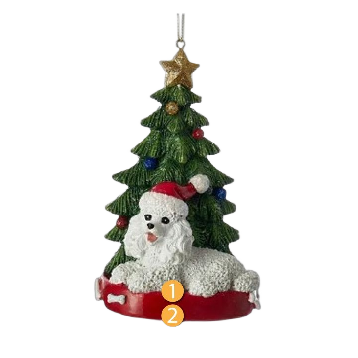 White poodle figurine wearing red Santa hat lying on red base in front of decorated green Christmas tree ornament