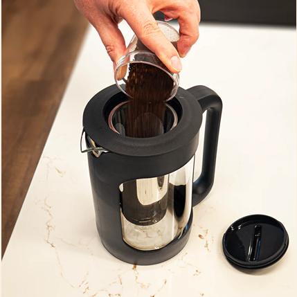 Hand pouring coffee grounds into black and clear Escali cold brew immersion coffee maker on white countertop
