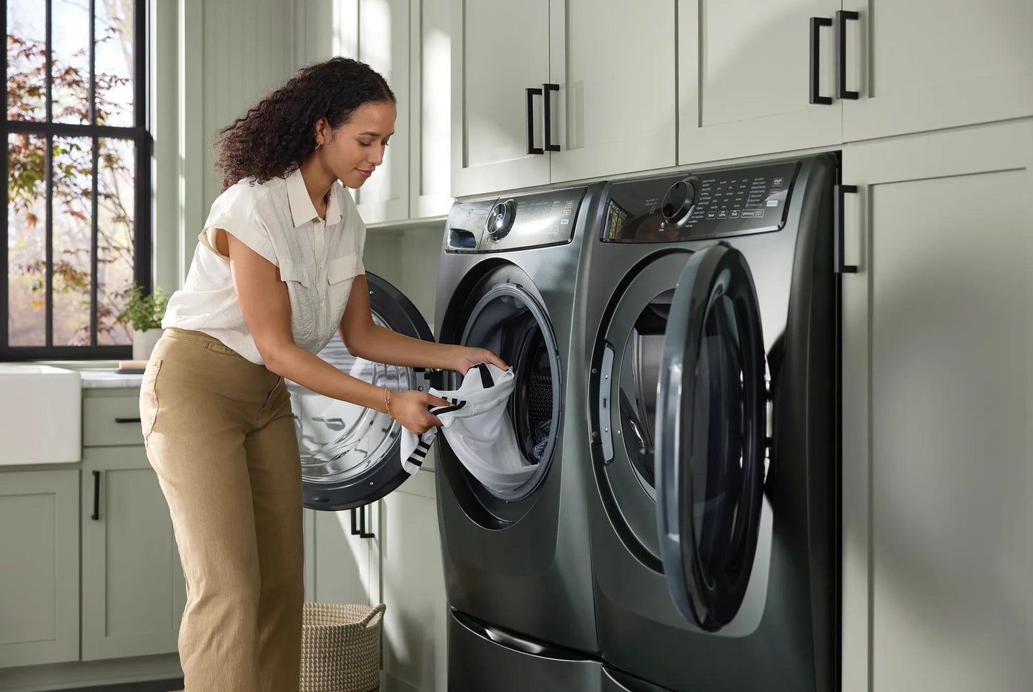 Woman placing laundry into dark gray front load washer in modern laundry room with light gray cabinets and large window