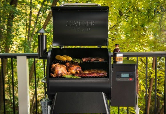 Outdoor black vertical smoker grill with cooked meats and corn inside, set on a porch railing surrounded by green leafy trees in the background.