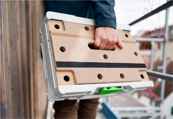Close-up of a person carrying a compact, beige and white plastic storage case with a green latch on a construction site background.
