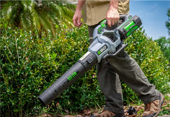 Person holding a green and black cordless battery-operated leaf blower in a garden with dense greenery and trees in the background.