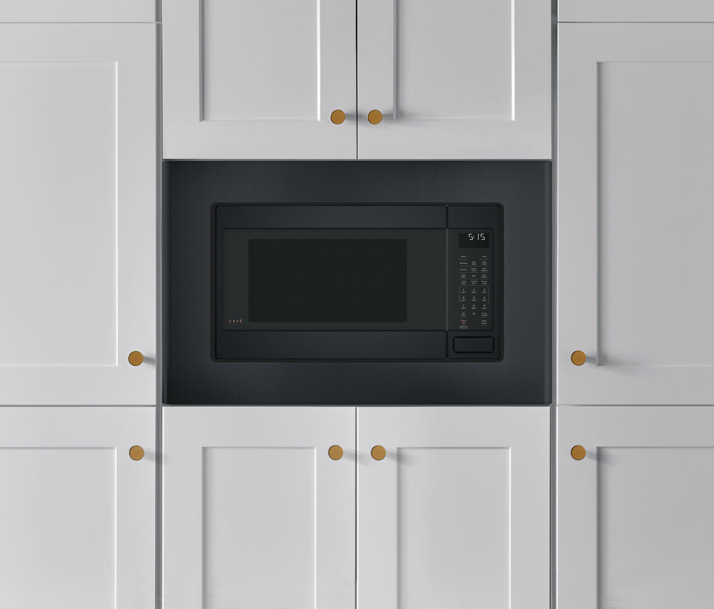 Matte black built-in trim kit surrounding a microwave installed in white kitchen cabinetry with brass hardware handles