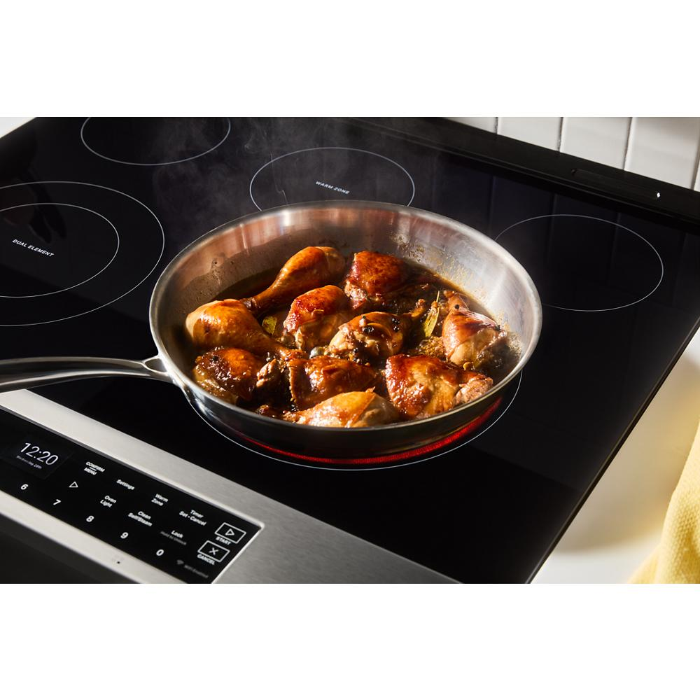 Stainless steel frying pan with chicken pieces cooking on smooth black glass electric induction cooktop surface