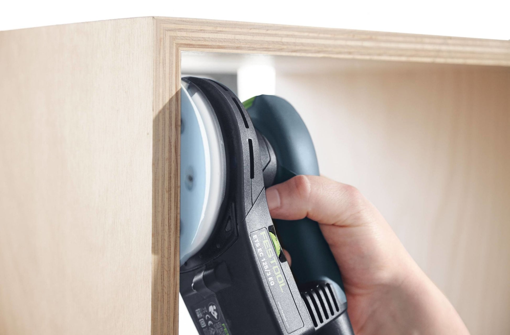 Hand holding a Festool random orbit sander smoothing the inside edge of a wooden cabinet corner