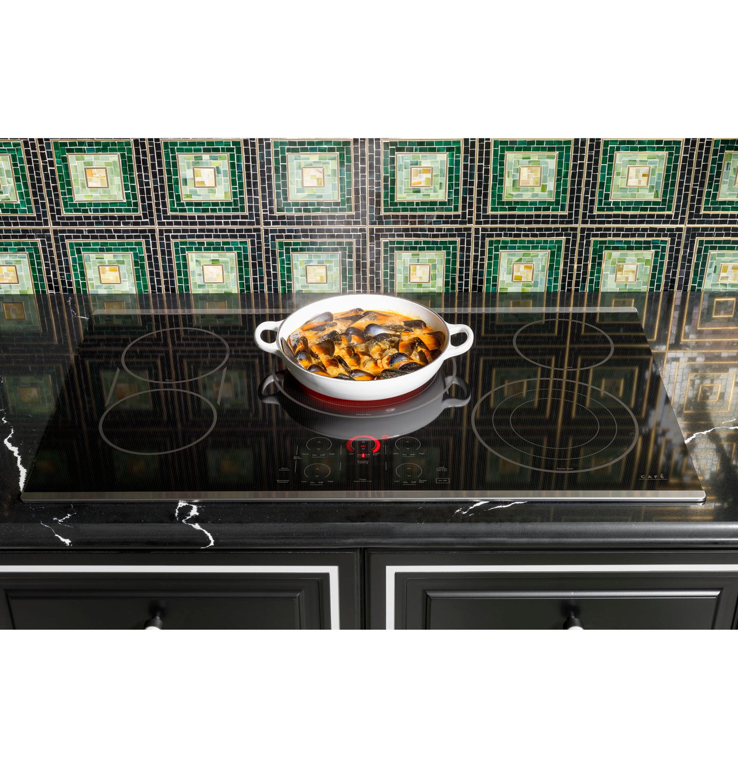 Red and white ceramic pot cooking food on black glass induction cooktop with green tile backsplash in kitchen