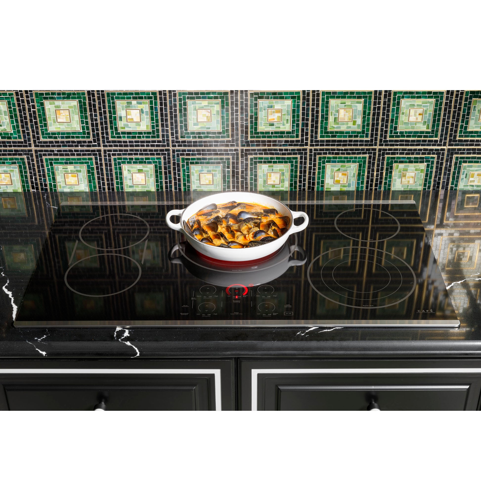 Red and white ceramic pot cooking food on black glass induction cooktop with green tile backsplash in kitchen