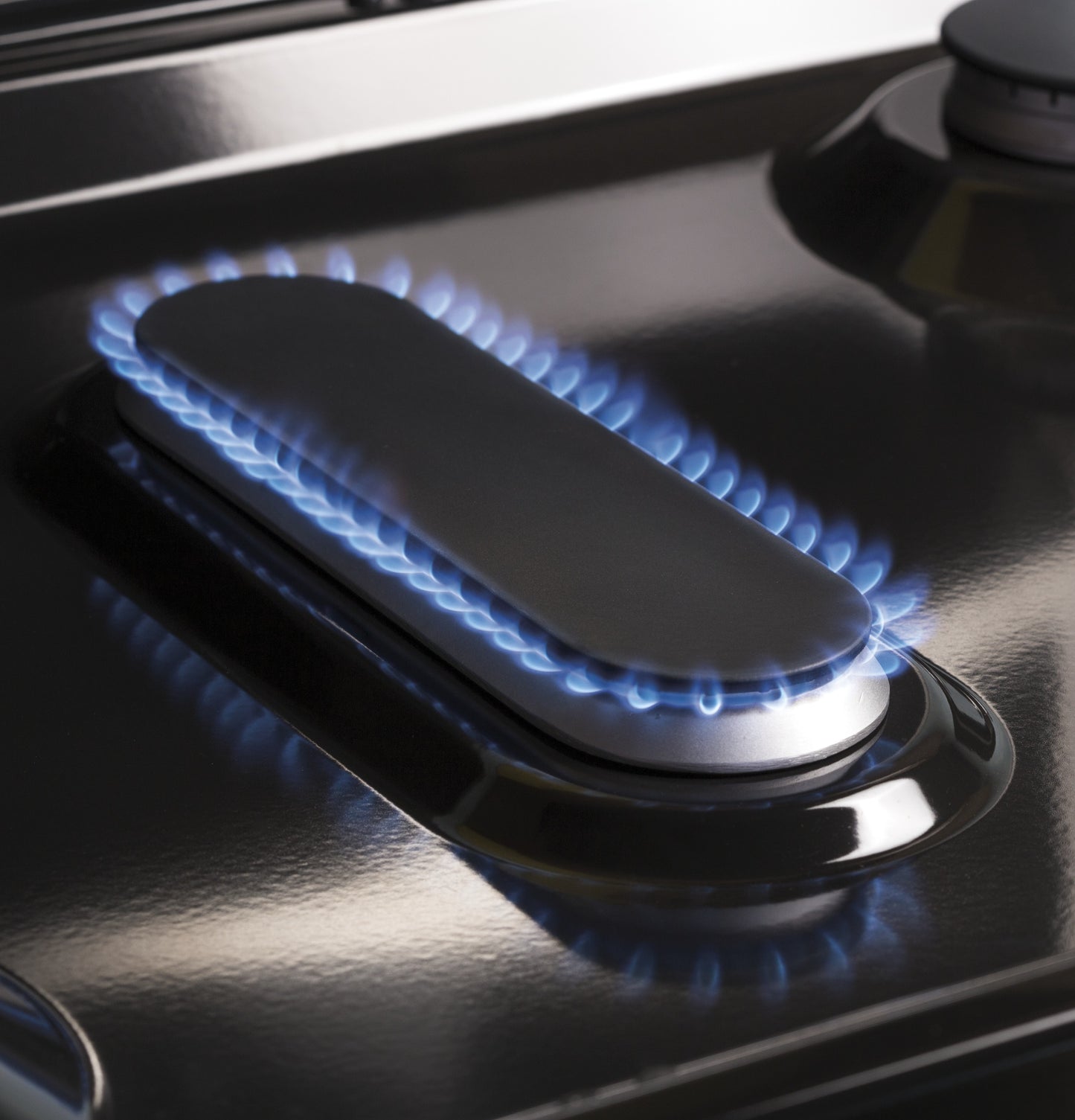 Close-up of a blue flame burning around an elongated burner on a black gas range cooktop for home appliances.