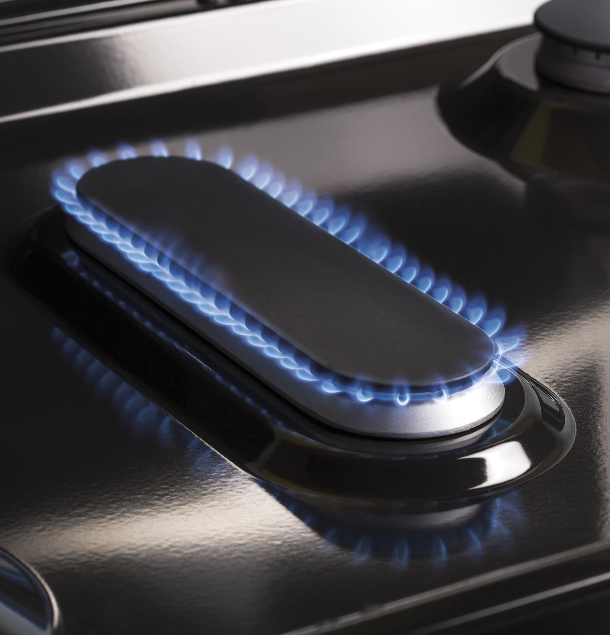 Close-up of a blue flame burning around an elongated burner on a black gas range cooktop for home appliances.