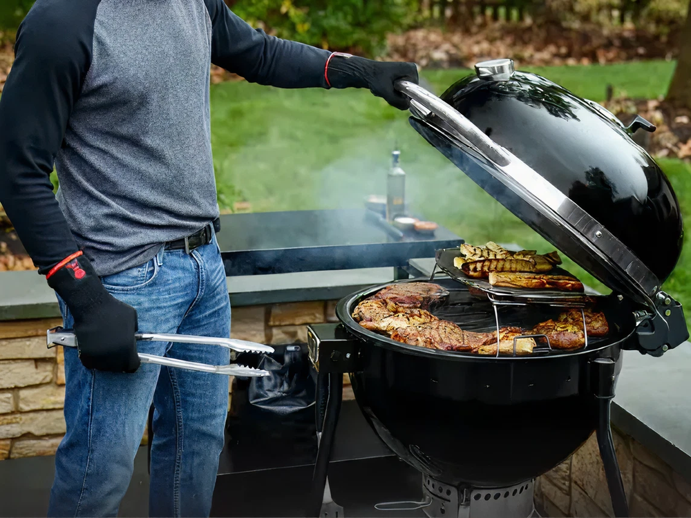 Person wearing gloves using tongs to lift the lid of a charcoal grill filled with various grilled meats and vegetables in an outdoor patio setting.