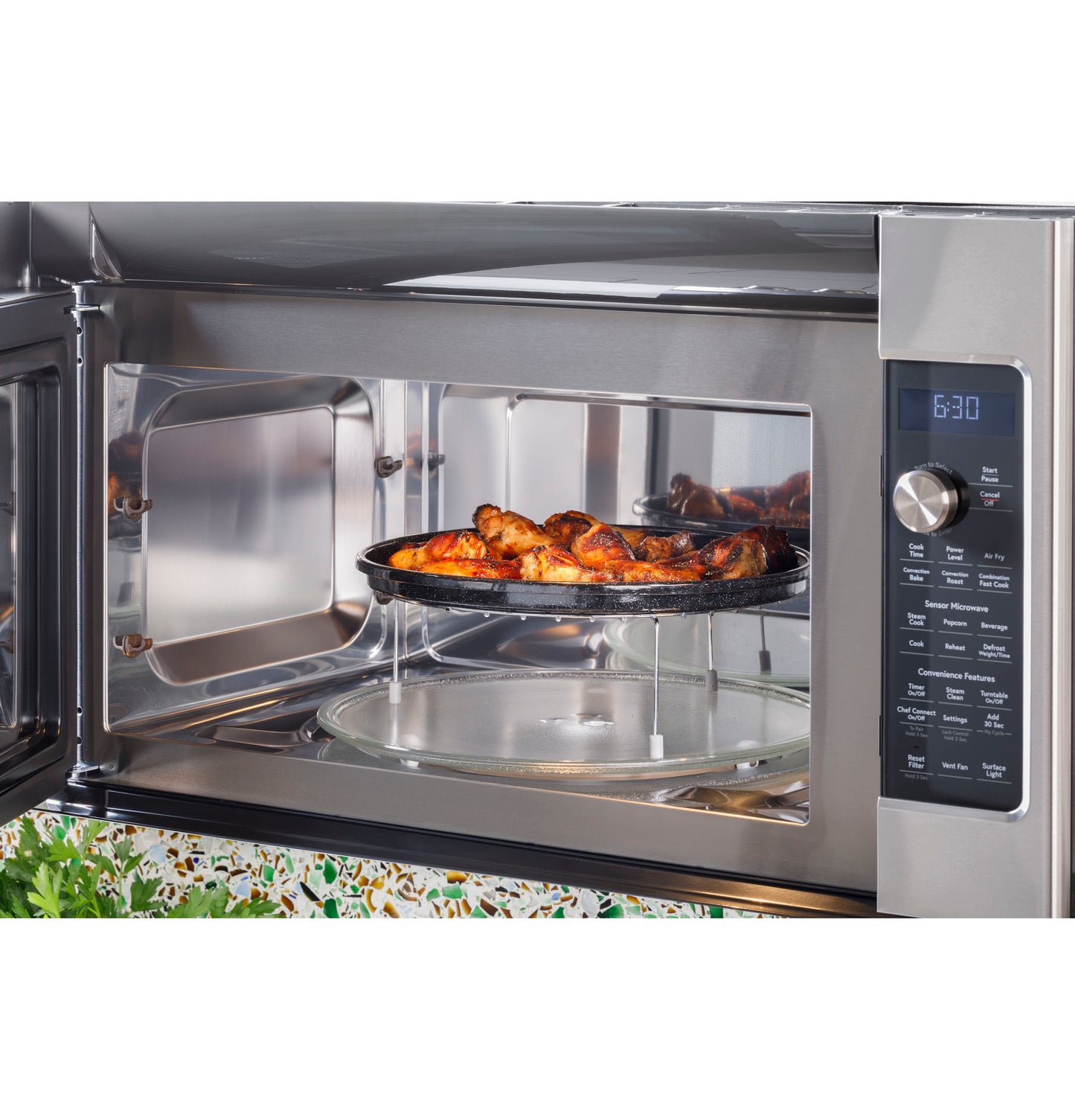 Stainless steel over-the-range convection microwave with open door showing cooked chicken on elevated tray inside kitchen appliance