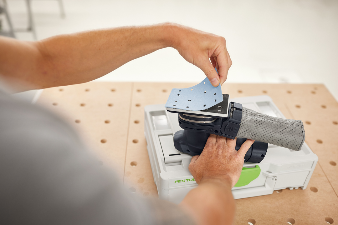 Hand placing blue abrasive sanding sheet onto a gray Festool sander for tool accessories and home improvement