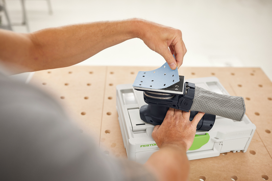 Hand placing blue abrasive sanding sheet onto a gray Festool sander for tool accessories and home improvement