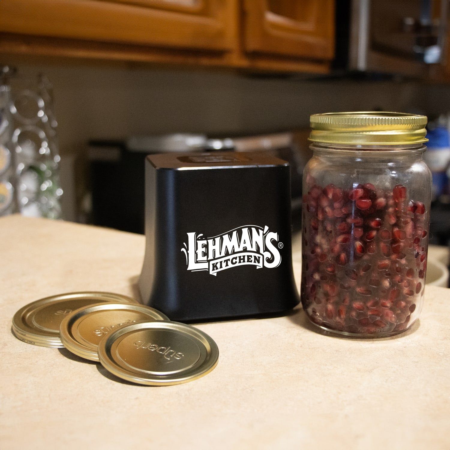 Black cordless vacuum sealer with three gold lids and glass jar filled with pomegranate seeds, kitchenware collection