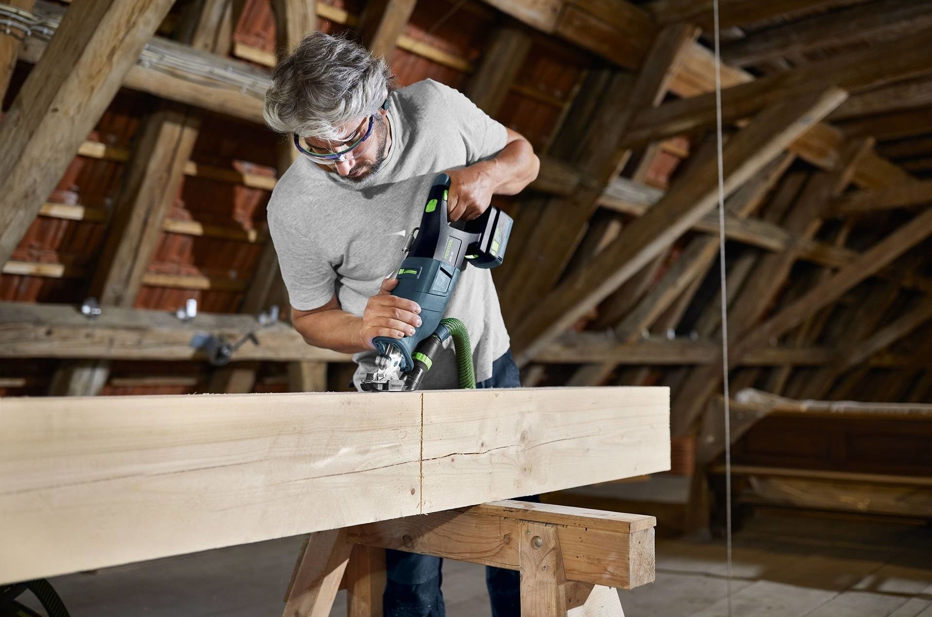 Man using cordless reciprocating saw on large wood beam in rustic indoor workshop with exposed beams