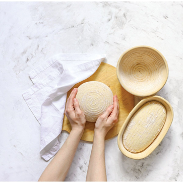 Hands holding round dough next to oval and round natural fiber bread proofing baskets with cloth liner on board in kitchenware collection