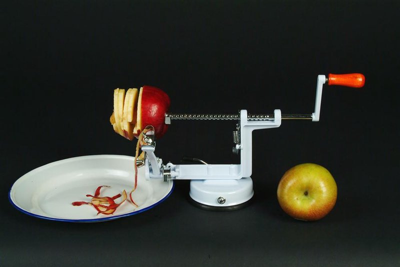 White and orange apple peeler with suction base peeling a red apple next to a whole yellow apple on black surface
