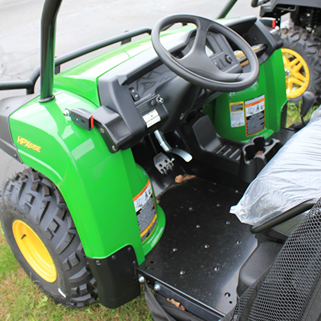 Interior view of green utility vehicle showing steering wheel, pedals, and rugged off-road tire on grassy ground