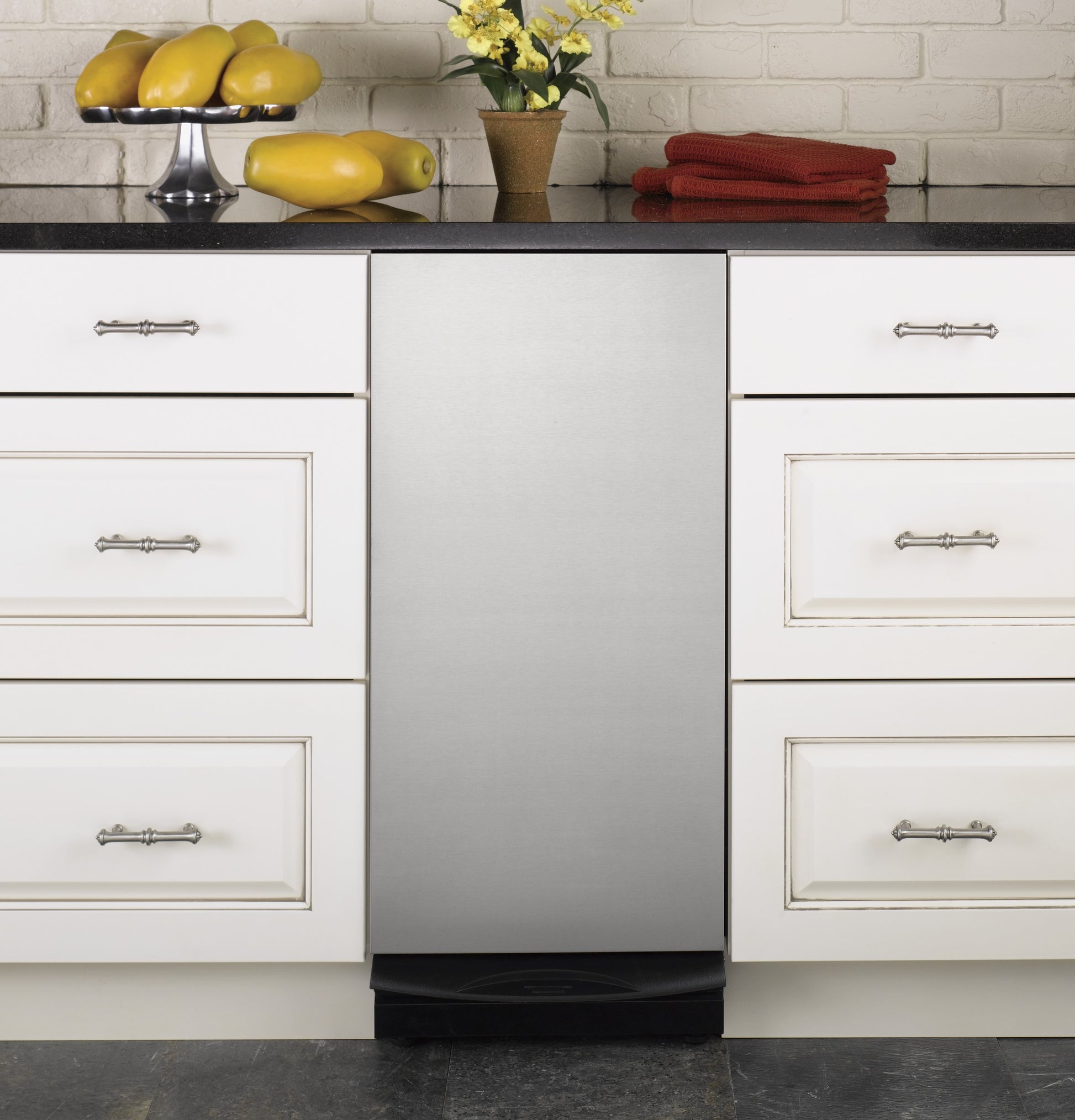 Built-in 15-inch stainless steel trash compactor integrated into white kitchen cabinetry with black countertop and decor items
