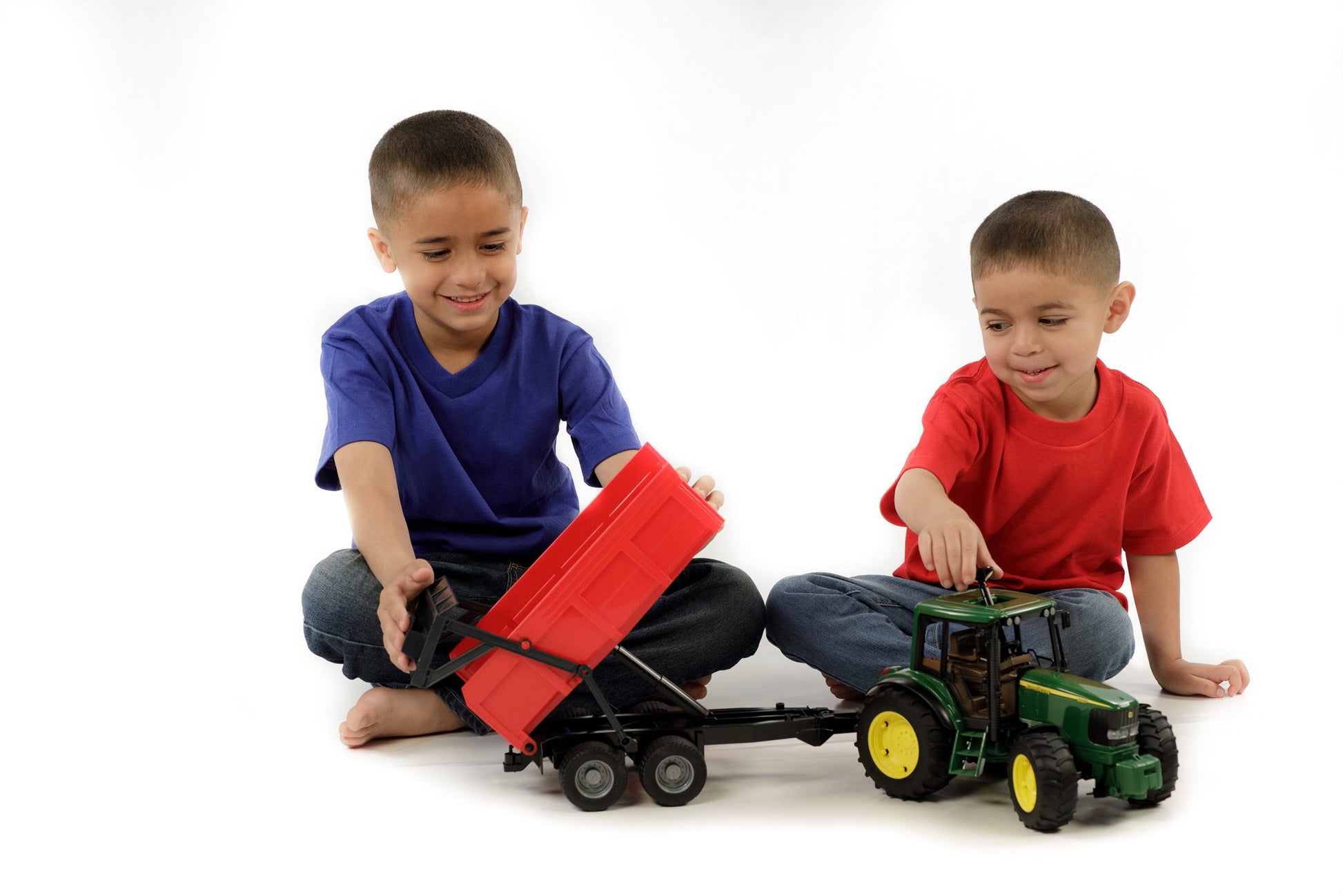 Two young boys playing with a detailed green toy tractor and red tipping trailer on a white background
