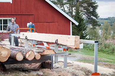 Man operating a Logosol F2 chain sawmill with dual log lifts cutting lumber outdoors near a red wooden building