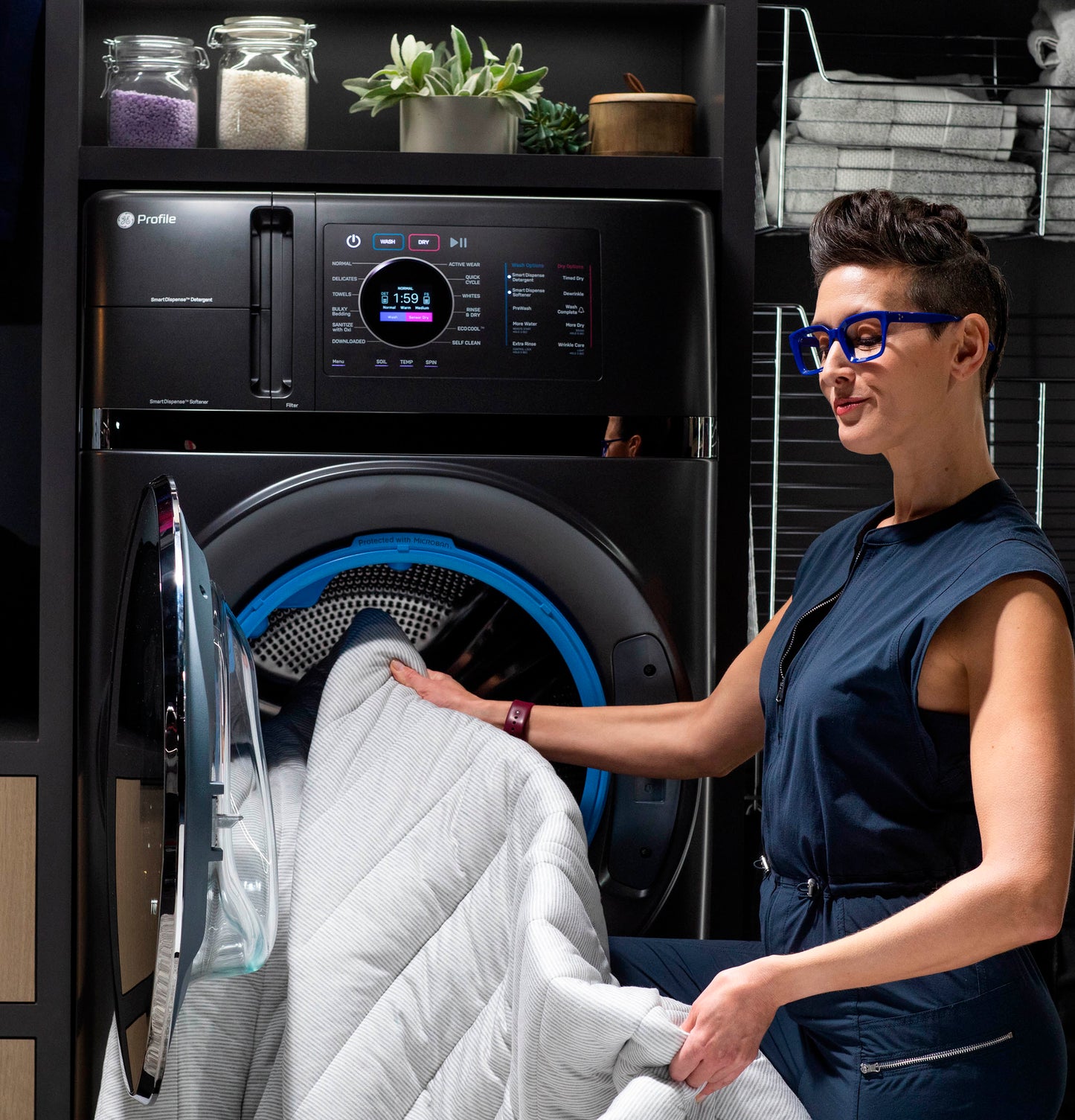Woman loading light gray quilt into black GE Profile washer/dryer combo in modern laundry space with shelves and towels