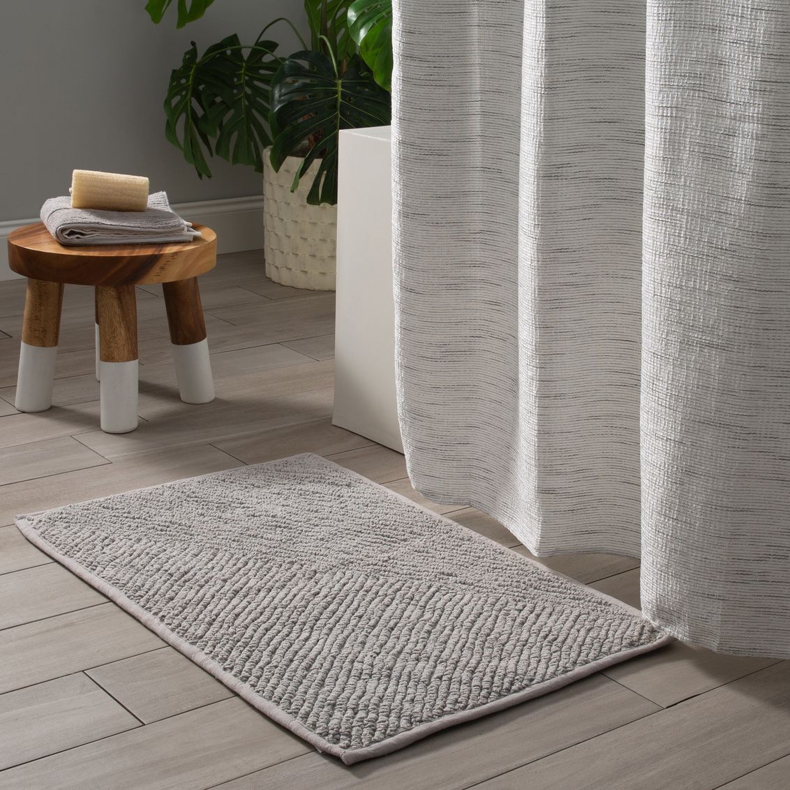 Gray textured bath mat with plush soft surface placed on wood bathroom floor near white curtain and wooden stool