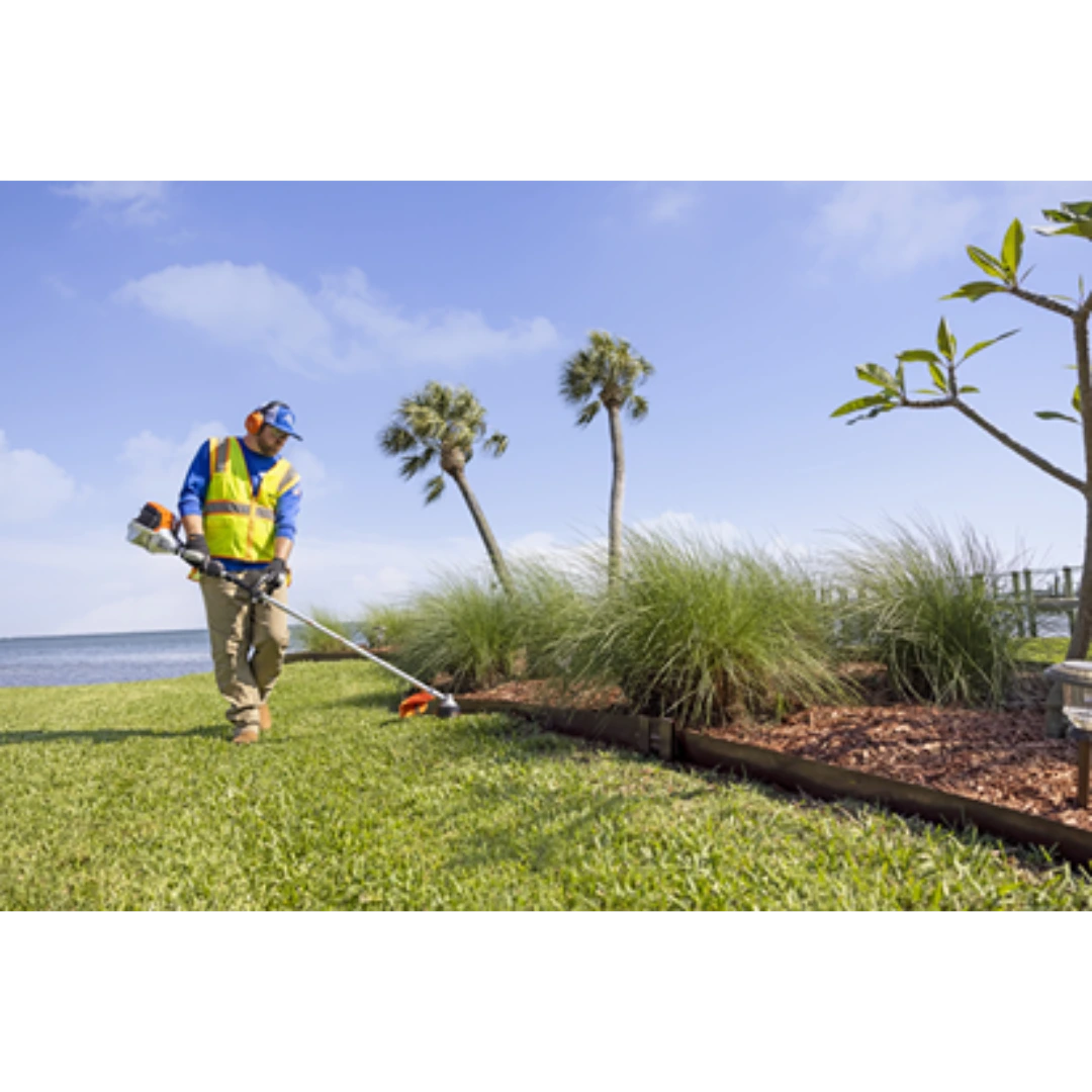 Worker using a loop handle string trimmer to edge grass along a landscaped garden bed near a water body