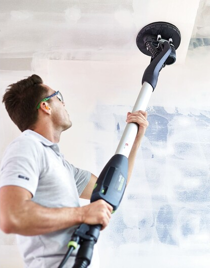 Man using a Festool drywall sander with an extended handle performing ceiling sanding in home improvement setting