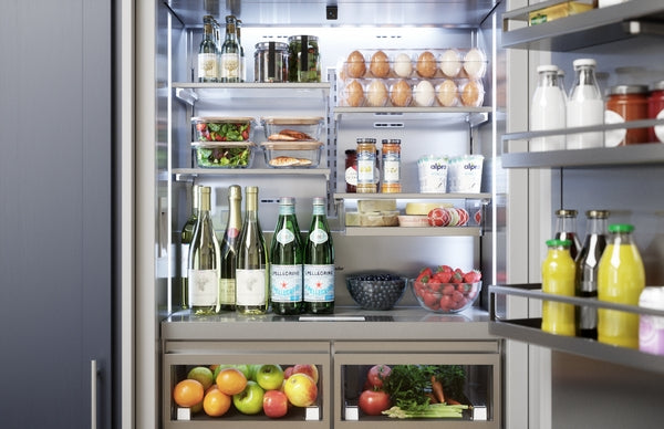 Open stainless steel refrigerator with organized shelves holding beverages, fresh produce, eggs, and condiments in kitchen appliance setting