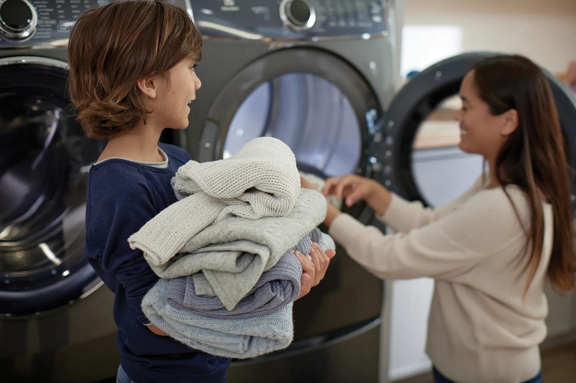 Child holding folded clothes near dark gray front load washer with woman loading laundry in background in home laundry room